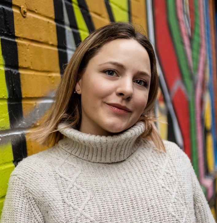 Image of Ettie. Ettie is a white person with light brown hair. She’s wearing a cosy beige jumper, and leaning against a colourful wall. She’s facing to the right, and smiling. Photo by the wonderful Sarah Tulej.