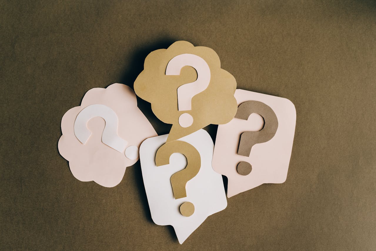 A set of four question marks crafted out of coloured paper and placed in the middle of speech and thought bubbles. The background is brown and the question marks and bubbles are a mix of lighter brown, light pink and white.